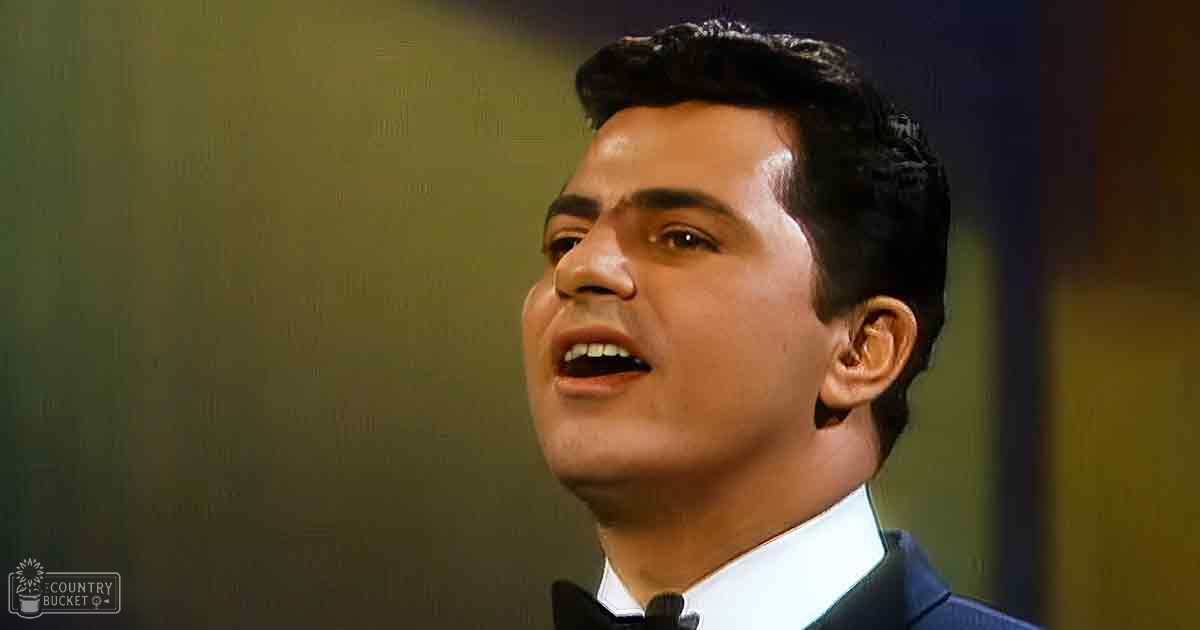 A young adult male in a well-fitted tuxedo stands against a plain, muted greenish wall.