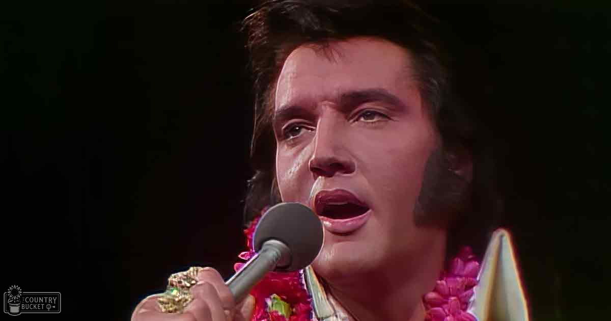An adult male performer in a stage setting, wearing a white mid-20th-century silk or satin garment with a high neckline and short sleeves, adorned with a pink floral lei around the neck. Holding an older-style microphone.