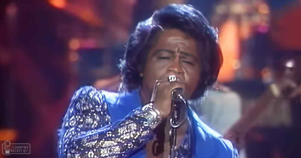 An adult female singer wearing a blue sequined 1970s-style garment with long sleeves and high neckline, holding a microphone at an indoor concert venue.