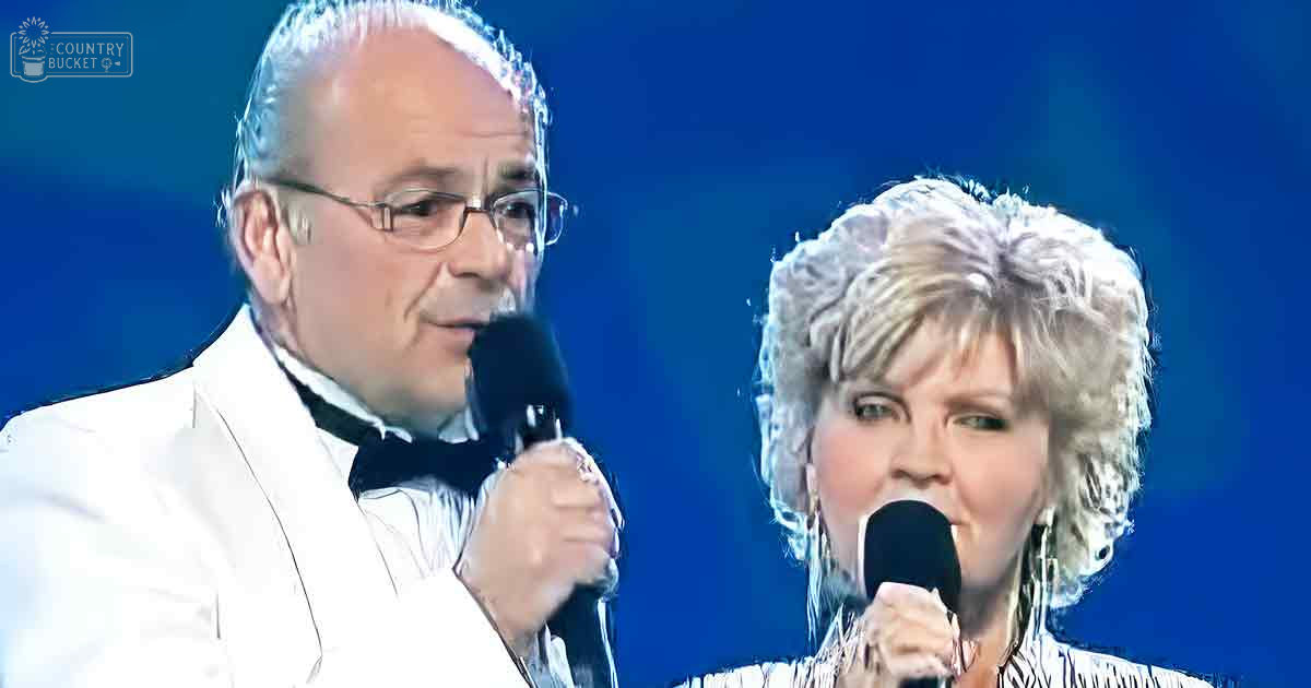 A middle-aged male singer in a formal white suit and bow tie performs alongside an older adult female in a light, textured top on stage under uniform blue lighting.