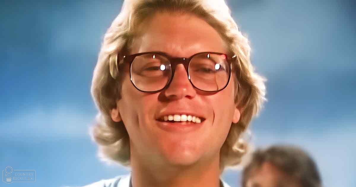 A young adult male with shoulder-length blonde hair, large round-framed glasses, and a light-colored collared shirt or jacket stands outdoors under a clear blue sky.