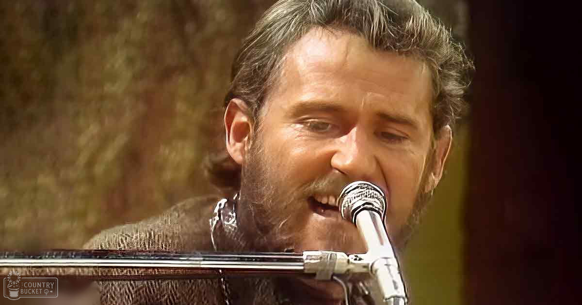 A middle-aged male singer wearing a textured brown winter coat with fur-lined collar, performing into a microphone on stage.