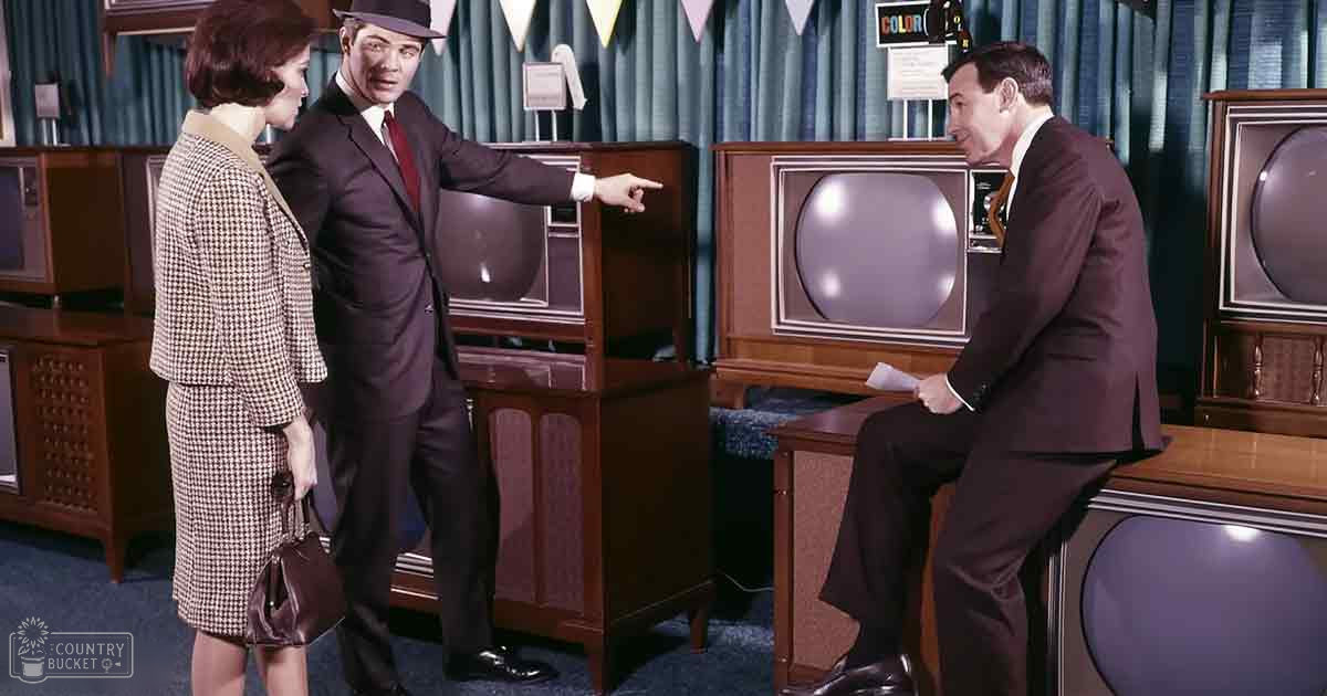 A man in a dark brown suit stands among vintage-style televisions and wooden stands, wearing black dress shoes; an adult female in a checkered dress with beige collar and cuffs holds a handbag, suggesting a mid-20th century exhibition or event setting.