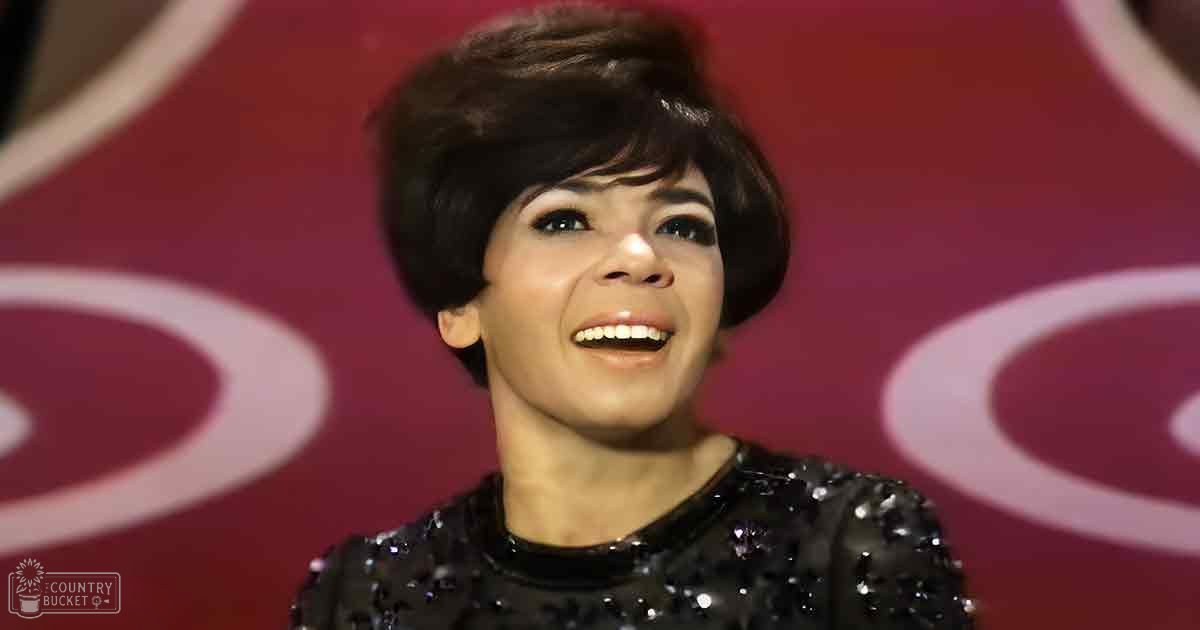 A young adult female in a form-fitting black sequined dress with reflective material, standing against a bright red backdrop with circular patterns.
