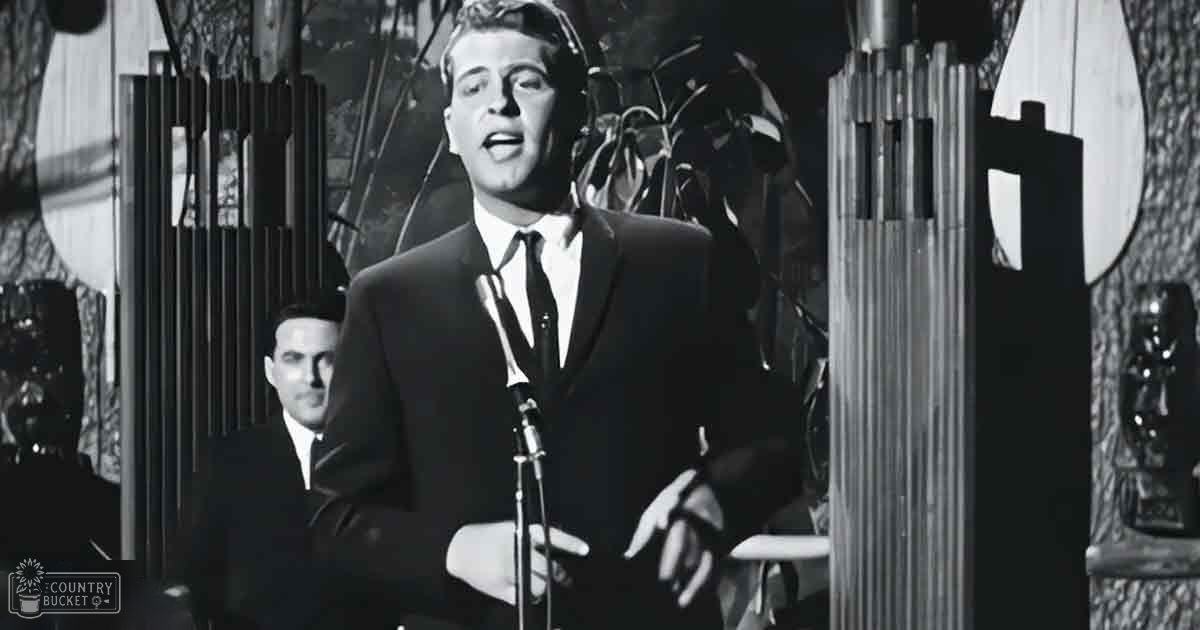 A young adult male singer in a dark suit with a white shirt and black tie performs on stage near a microphone; behind him, an ornate wall piece and plants create the ambiance of a mid-20th century performance venue.
