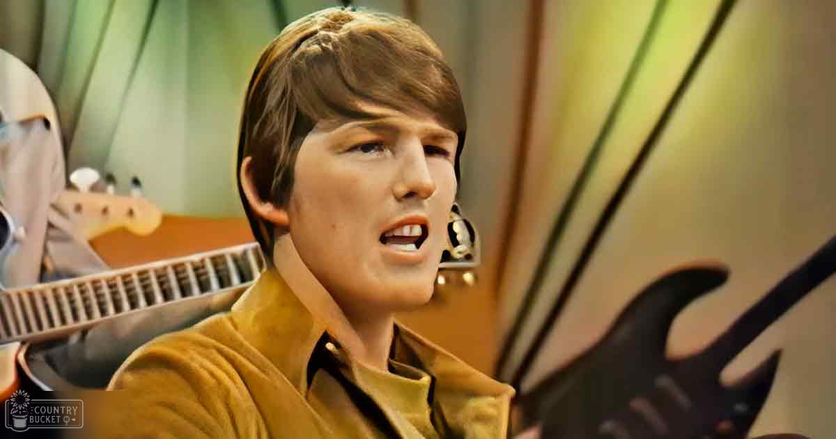 A young adult male wearing a mustard-yellow velour jacket with a high collar and long sleeves, surrounded by musical instruments in what appears to be a recording studio setting.