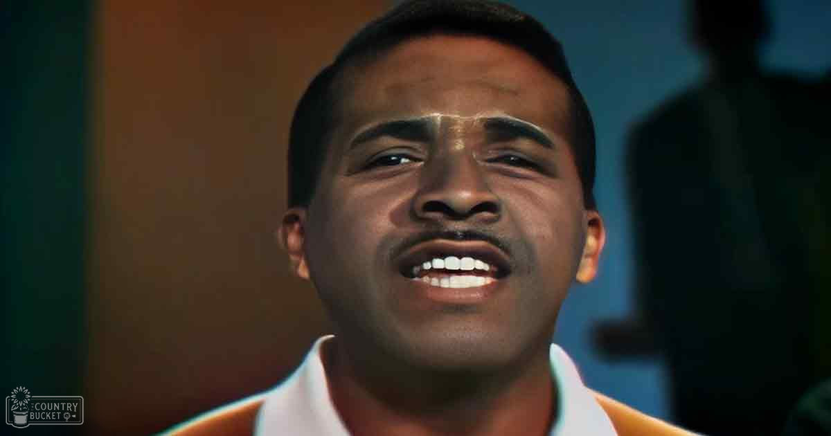 Young adult male in an orange collared shirt, singing indoors with dim lighting.