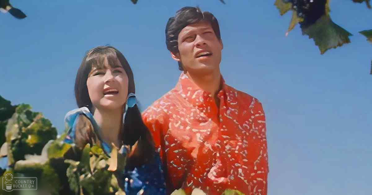 A young adult female and male wearing casual summer attire stand outdoors under clear skies, surrounded by green foliage.