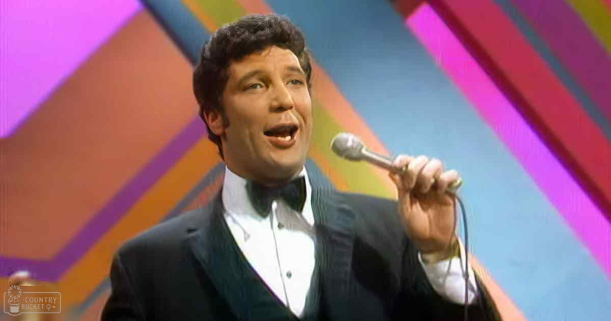A male adult performer in a dark suit jacket with a white shirt and bow tie on stage, holding a microphone against a vibrant geometric backdrop.