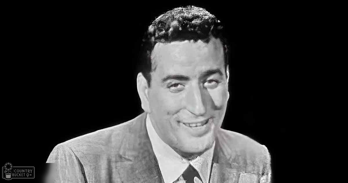 Adult male in a tailored dark suit with checkered jacket, standing indoors under stage or studio lighting.