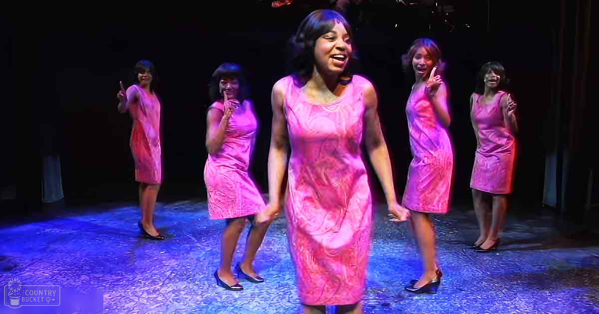 A young adult female in a theatrical performance setting, wearing a pink floral dress with lace trim and black high-heeled shoes; four other individuals are dressed similarly in the background.
