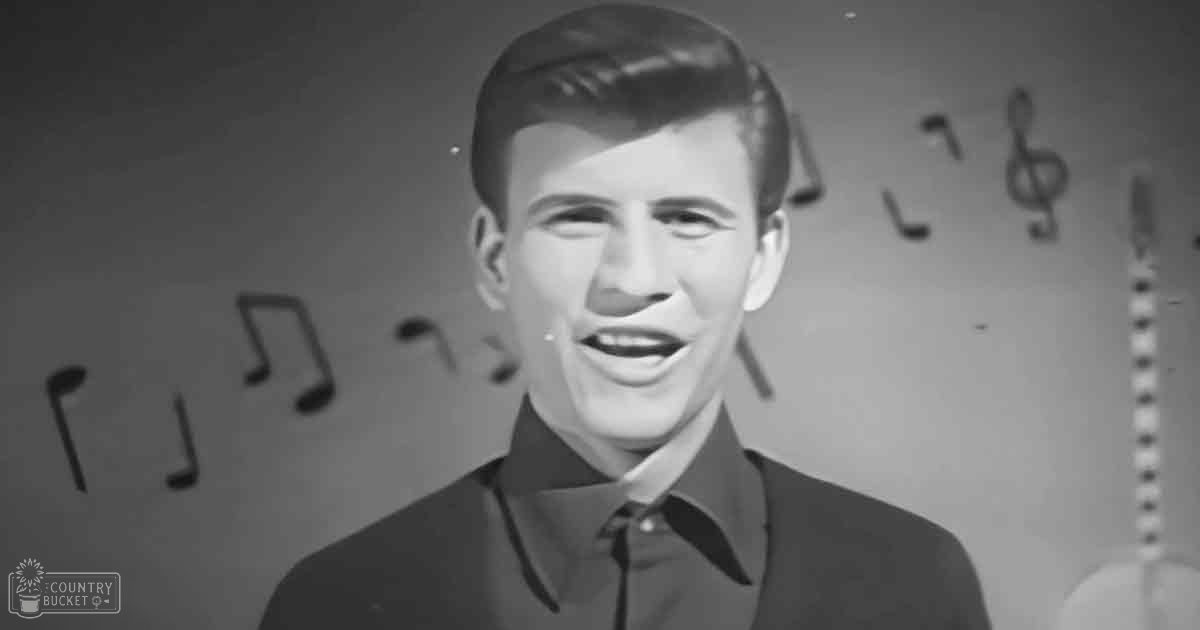 A young adult male wearing a dark-colored suit jacket and shirt with a tie, positioned indoors against a plain wall with musical notes, suggesting a formal or performance setting.