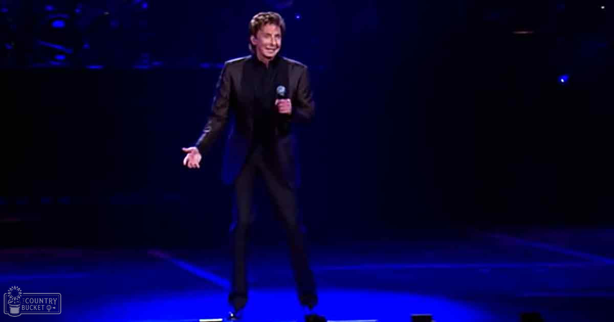 An adult male singer in a black suit jacket and dress pants performing on an indoor stage with blue stage lighting.