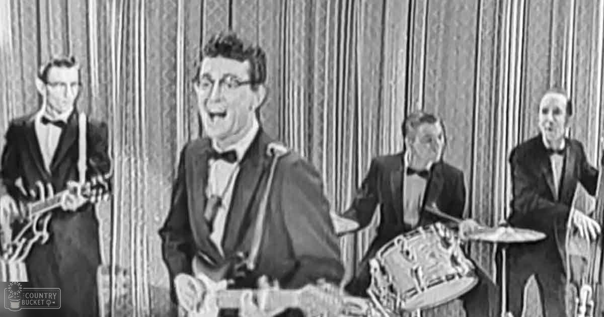 Young adult male musician wearing a dark suit with a bow tie, playing guitar in an indoor music performance setting with vertical blinds or curtains as backdrop.