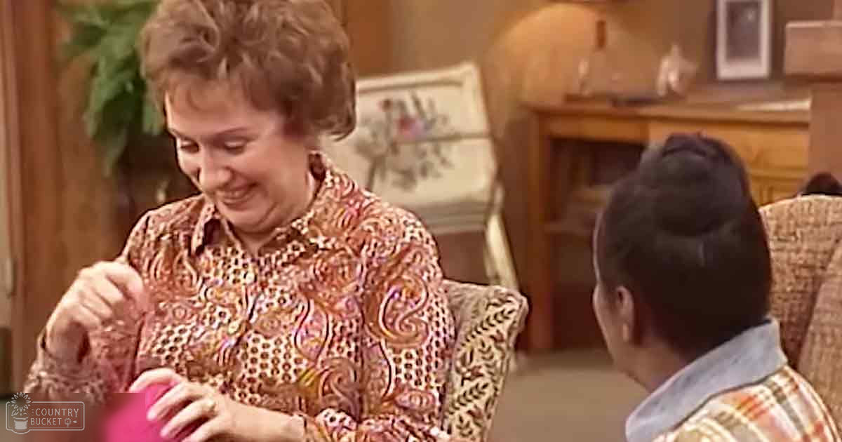 An older adult woman wearing a patterned blouse in a cozy living room setting with wicker furniture, fireplace, and framed artwork.