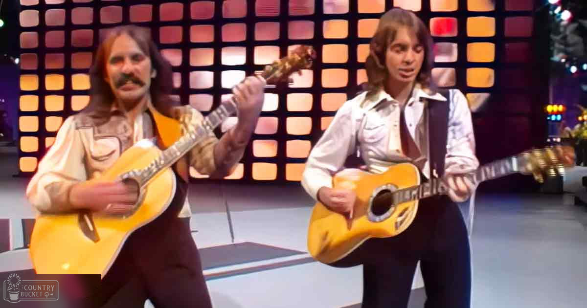 A musical performance scene with two adult male singers in an indoor stage setting under warm, retro-style lighting.