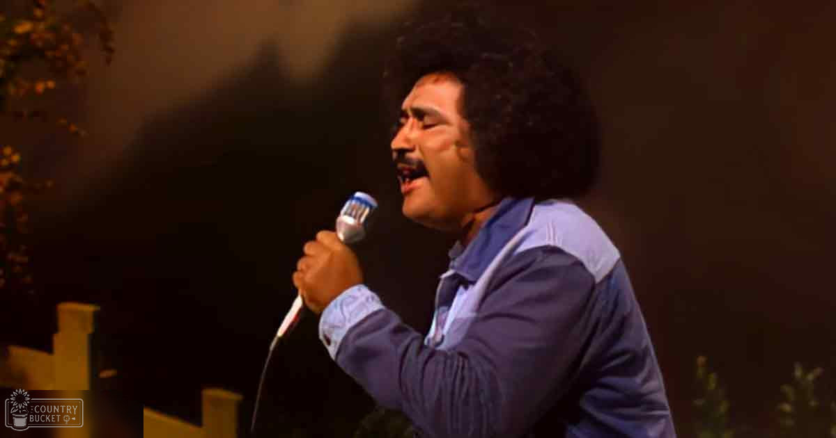 An adult male singer wearing a long-sleeved, collared shirt with a light blue and white patterned design performs on stage, holding a microphone against dimly lit walls with blurred greenery in the background.