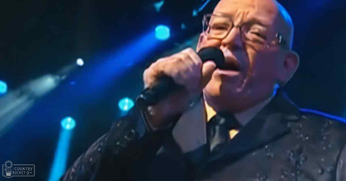 An older adult male singer wearing a dark, textured jacket with small studs or dots, singing into a microphone on an indoor stage illuminated by blue lighting.