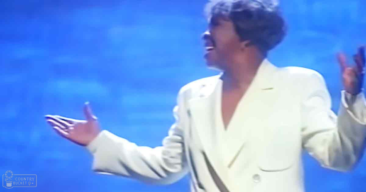 A middle-aged female performer wearing a white mid-20th century-style blazer with 'Madly Odd' text, standing against a blue backdrop in an indoor stage setting.