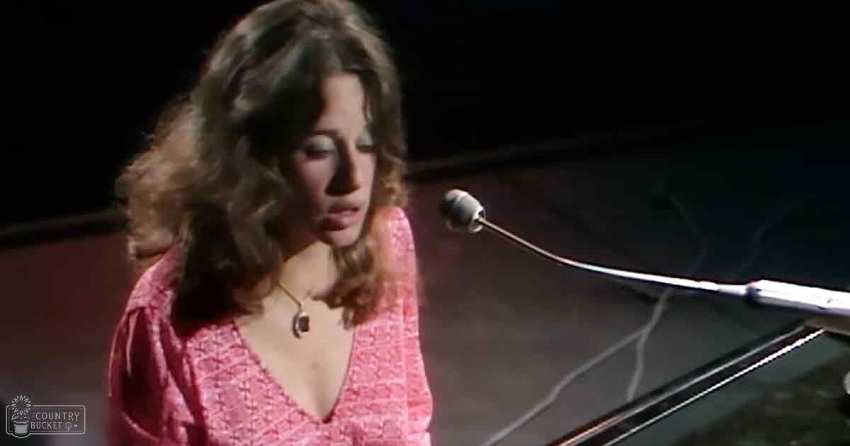 A young adult female in a 1970s-style pink patterned blouse performs at an indoor event, possibly singing into a microphone near a grand piano.