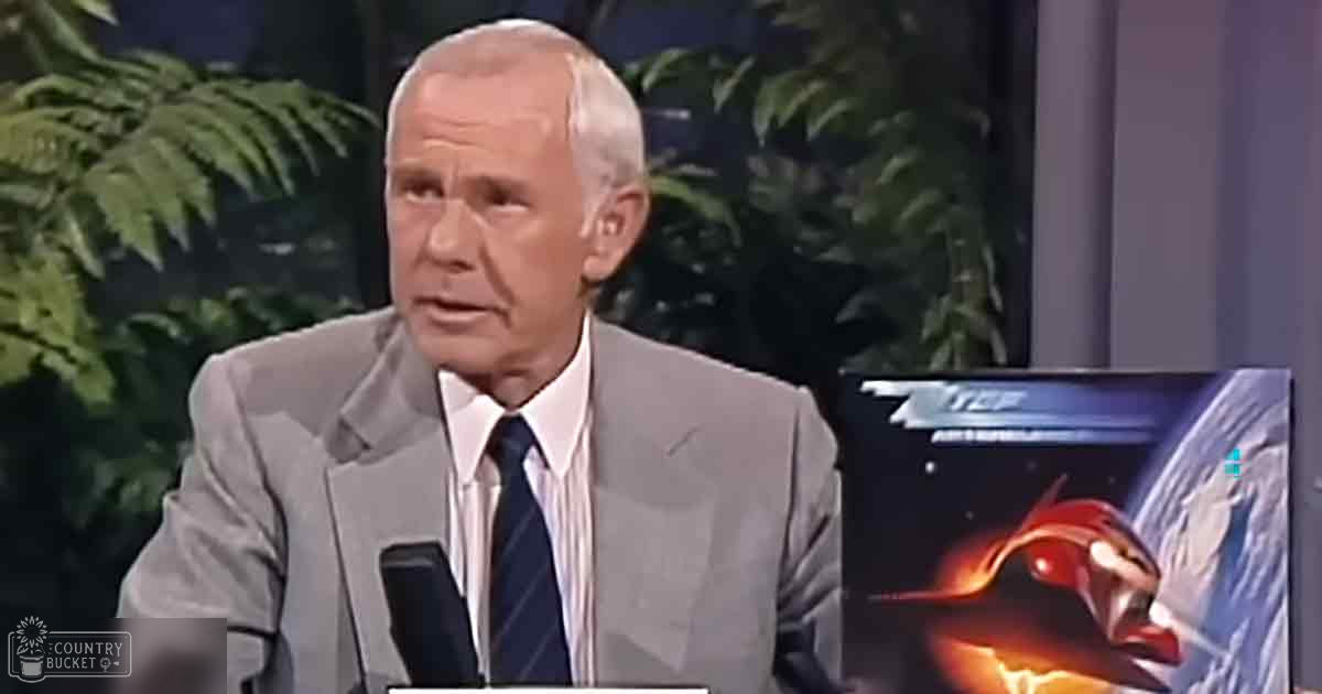 Middle-aged man in a tailored gray suit jacket with a white dress shirt and black tie, seated at a desk with a microphone and a backdrop suggesting a talk show environment.