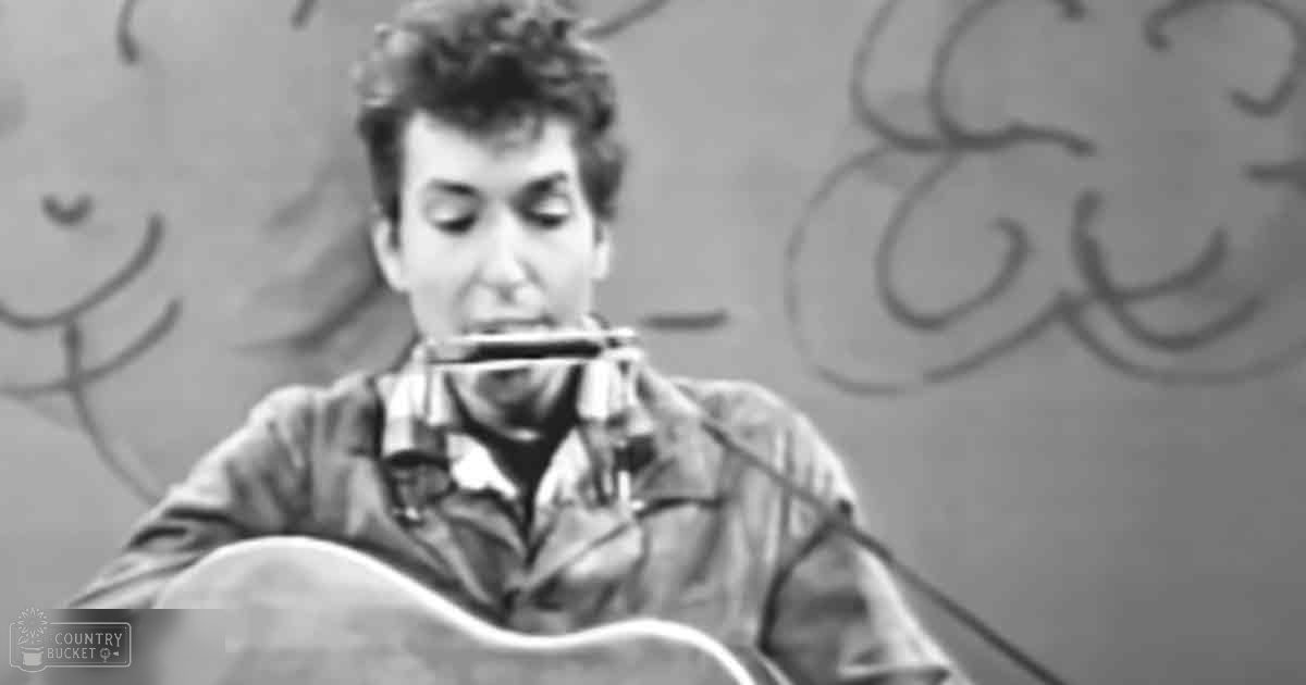 A young adult male wearing a plaid shirt and light-colored jacket plays harmonica in front of a microphone, set against an urban backdrop with graffiti art.