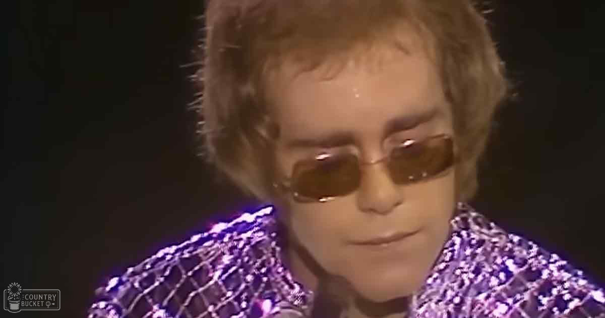 An adult male singer in a glittery, sequined purple jacket with white dot patterns and high collar indoors. Low lighting reflects off the shiny fabric.