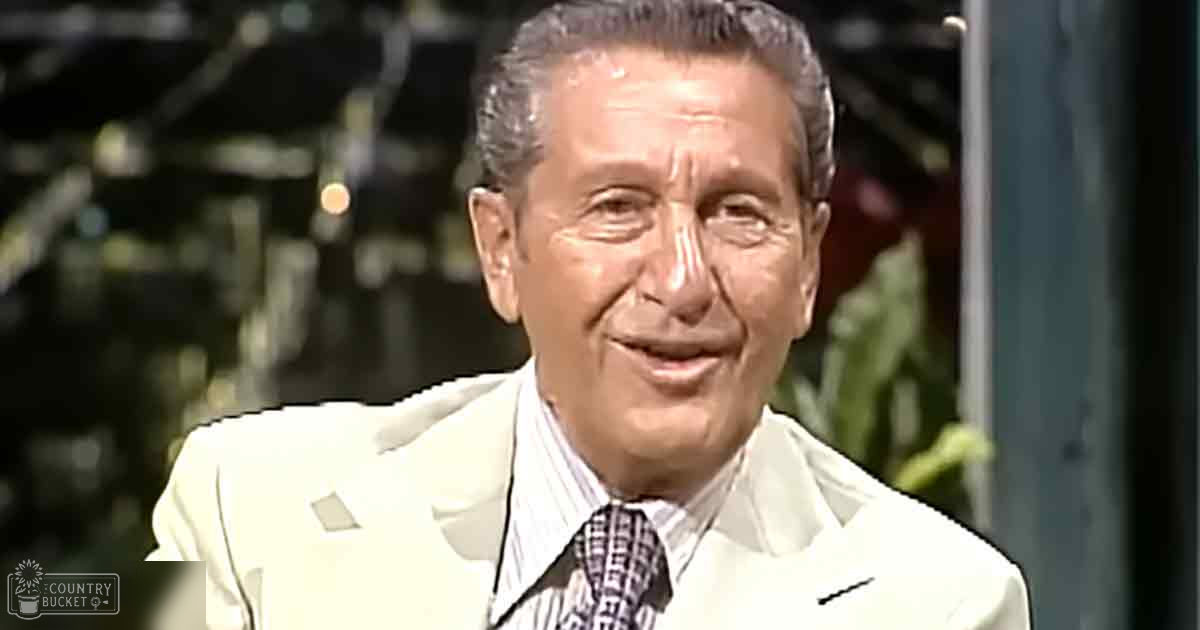 An older adult male dressed in a white suit jacket with subtle patterns, standing indoors near decorative greenery, possibly at an event or studio setting.