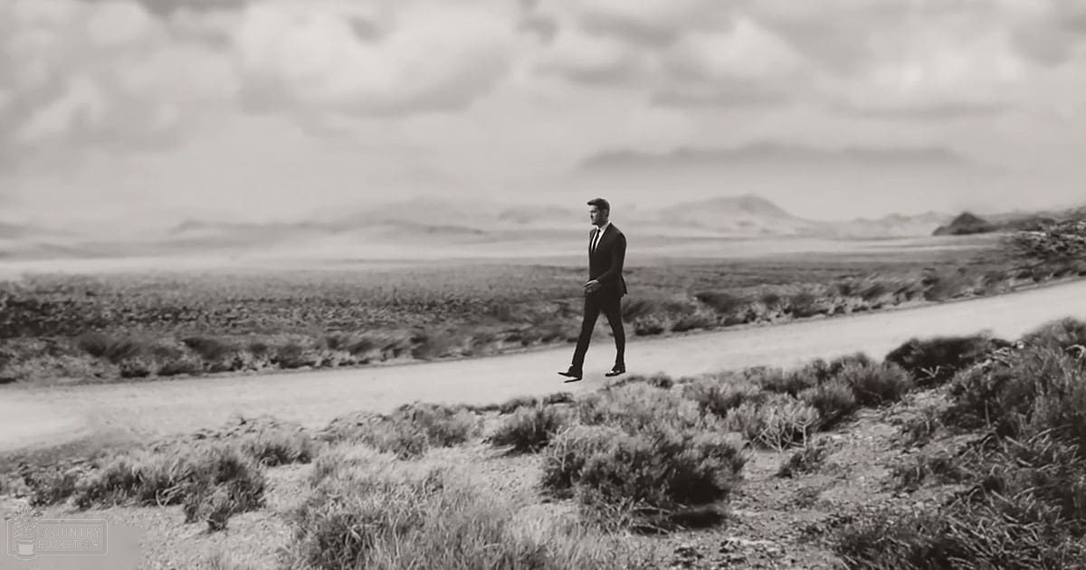 Adult male wearing a dark suit and tie walks across an arid, cloudy desert landscape with muted tones suggesting midday or early afternoon light.