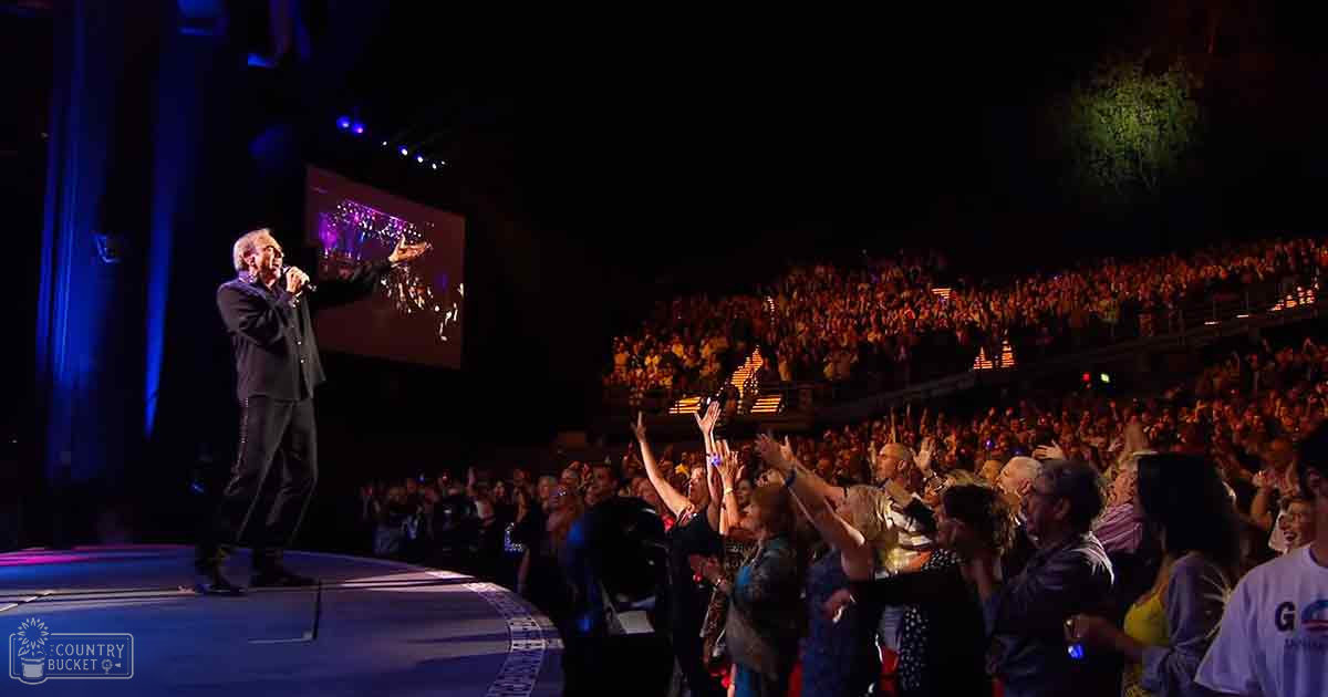 Adults of varying ages gather at an indoor concert venue, watching a performer on stage under spotlights; audience members wear casual or semi-formal attire with varied colors.