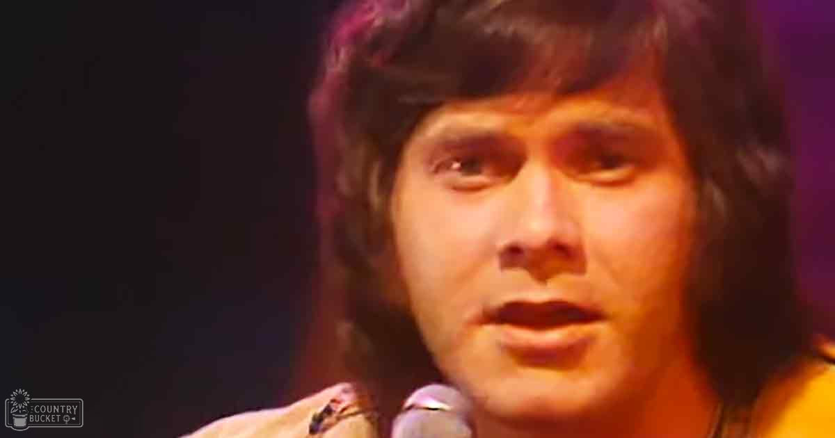 A young adult male in a patterned garment, possibly from the 1970s era, wearing a collar and long sleeves, standing on stage with a microphone under purple lighting.