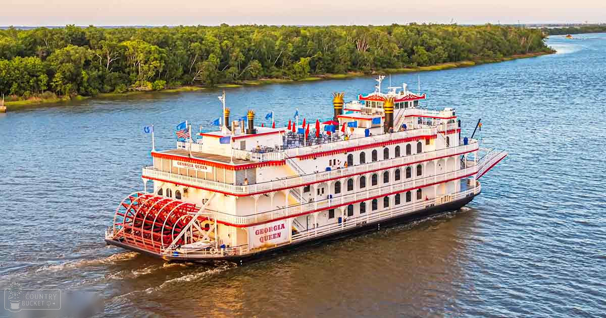 A serene river setting with a large white paddlewheel boat named 'Georgia Queen,' adorned with red accents, blue flags, and red umbrellas; lush green trees line the calm waters under soft golden evening light.