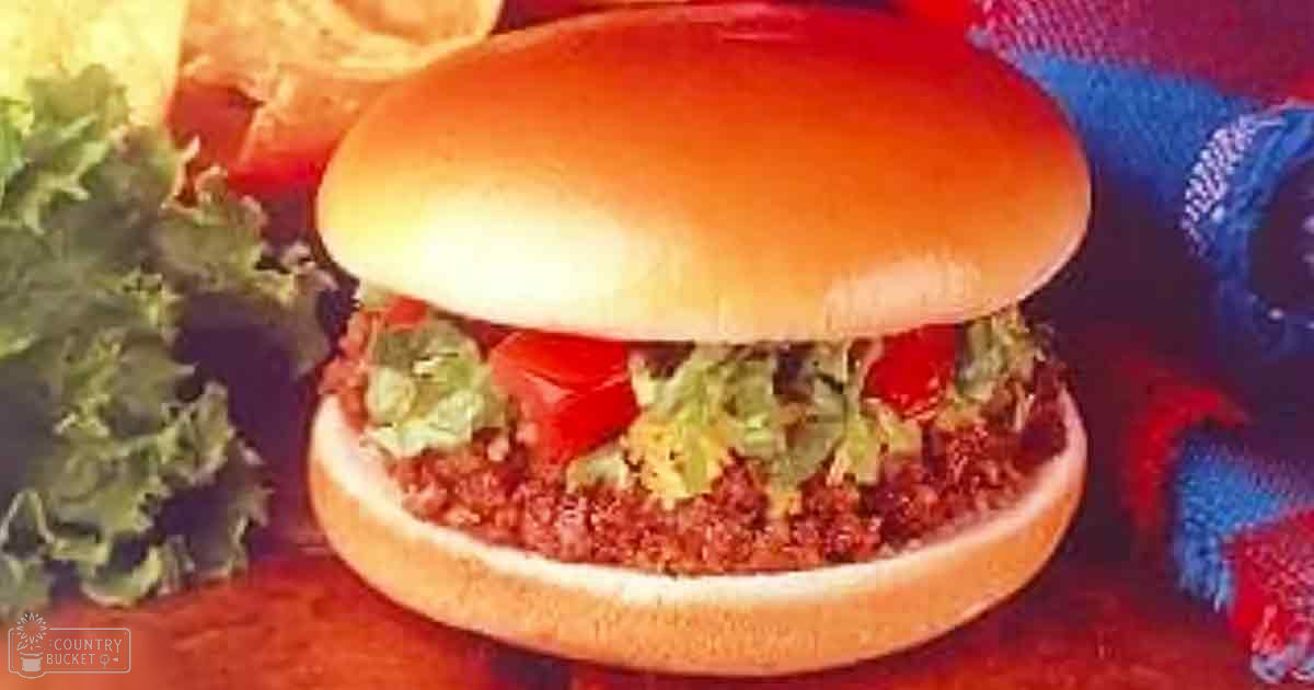 A plated hamburger with lettuce, tomato slices, and chips on a bun, set against a red and blue patterned cloth in an indoor casual dining setting