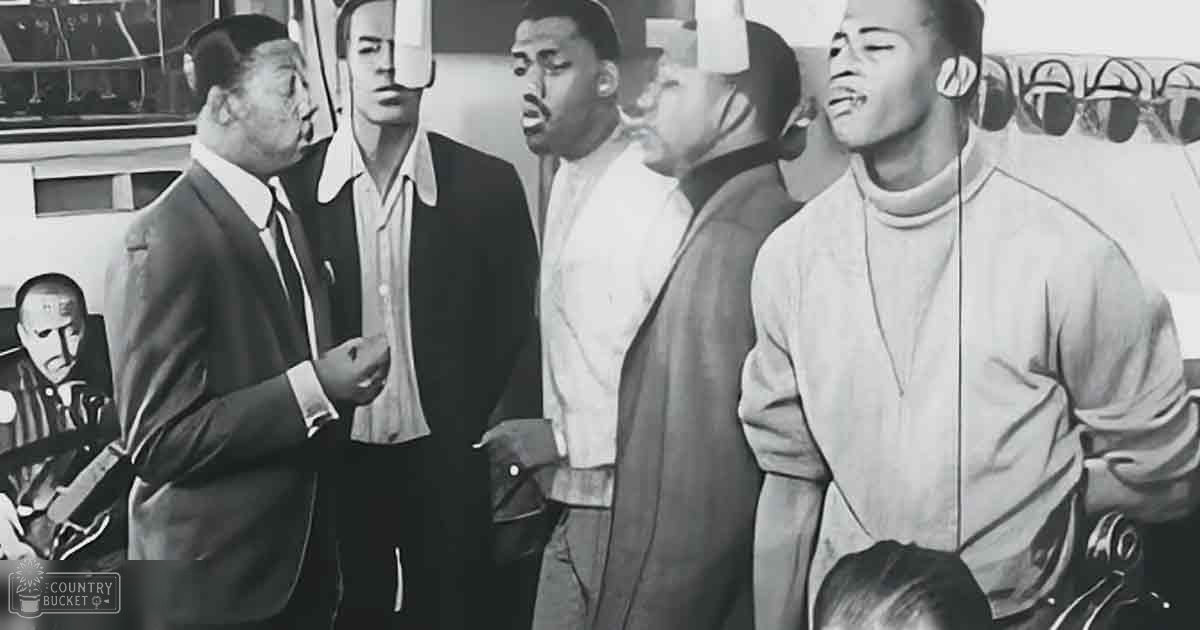 A group of individuals in casual to formal attire from the mid-20th century gather outdoors near what appears to be a waterfront, with railings visible in the background.