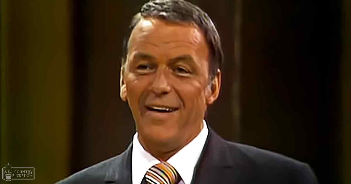 An older adult male wearing a dark suit jacket, white dress shirt, and striped tie stands in a well-lit indoor setting with dark curtains.