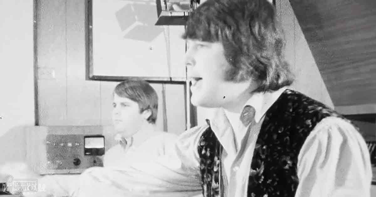 A young adult male stands in a recording studio, holding a microphone stand close to his mouth while another child or teenager looks on in the background. The setting includes an old-fashioned audio mixing console and framed artwork.
