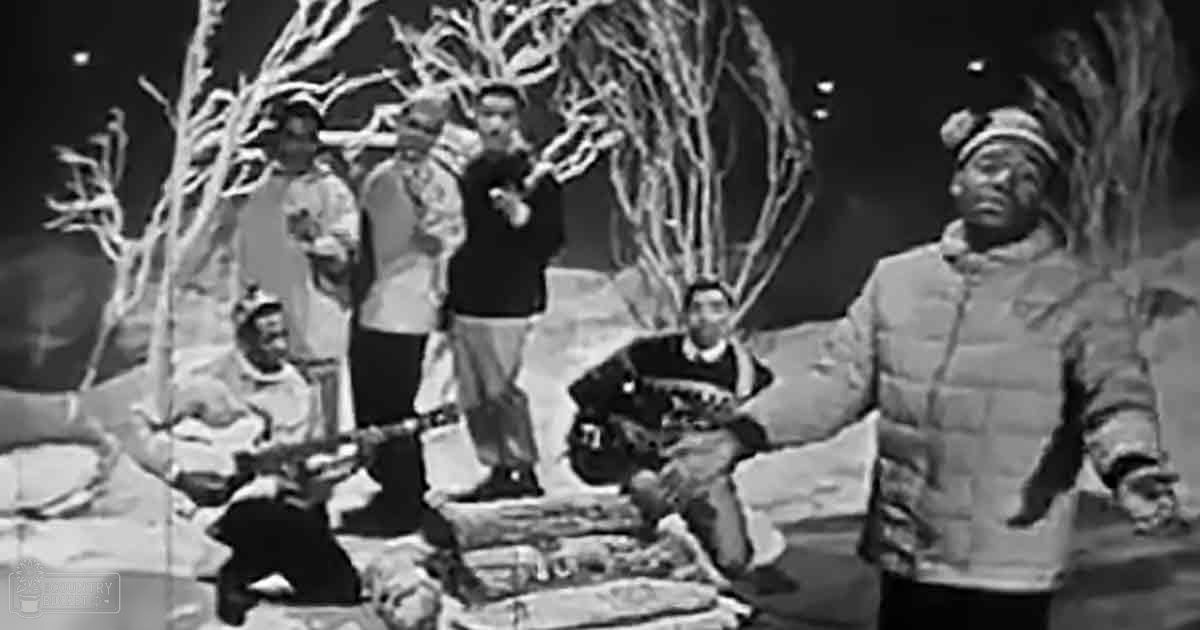 A group of individuals in a historical or dramatic outdoor winter scene with bare trees and snow-covered ground, possibly from a film or theatrical production.