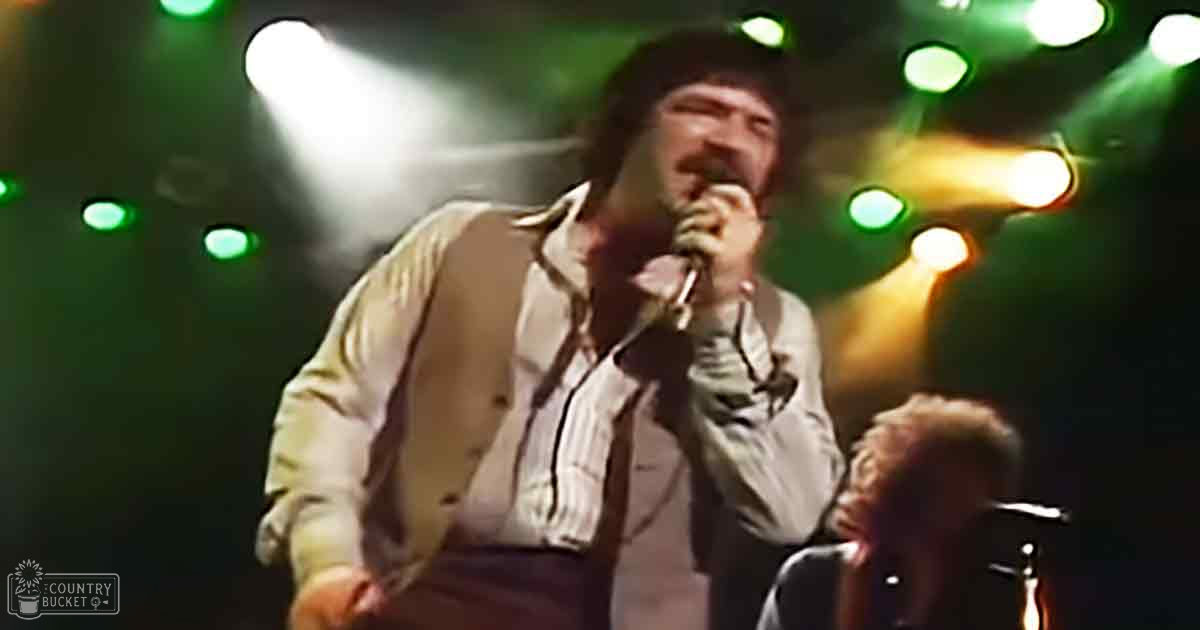 A male adult singer wearing a light beige button-down shirt and dark pants performs on stage at a concert venue under dim lighting, surrounded by stage lights.