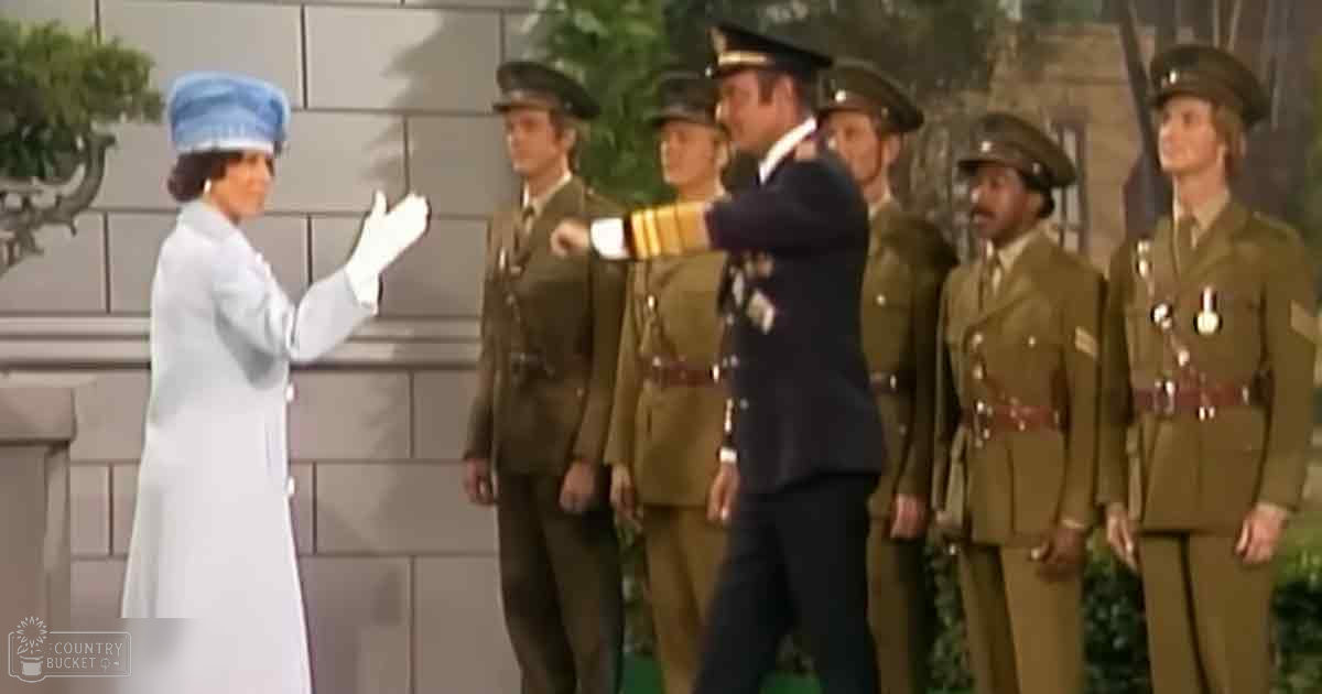 An adult female wearing a white coat dress with long sleeves and a high collar, accompanied by men in military uniforms outdoors near a building with greenery under natural daylight.