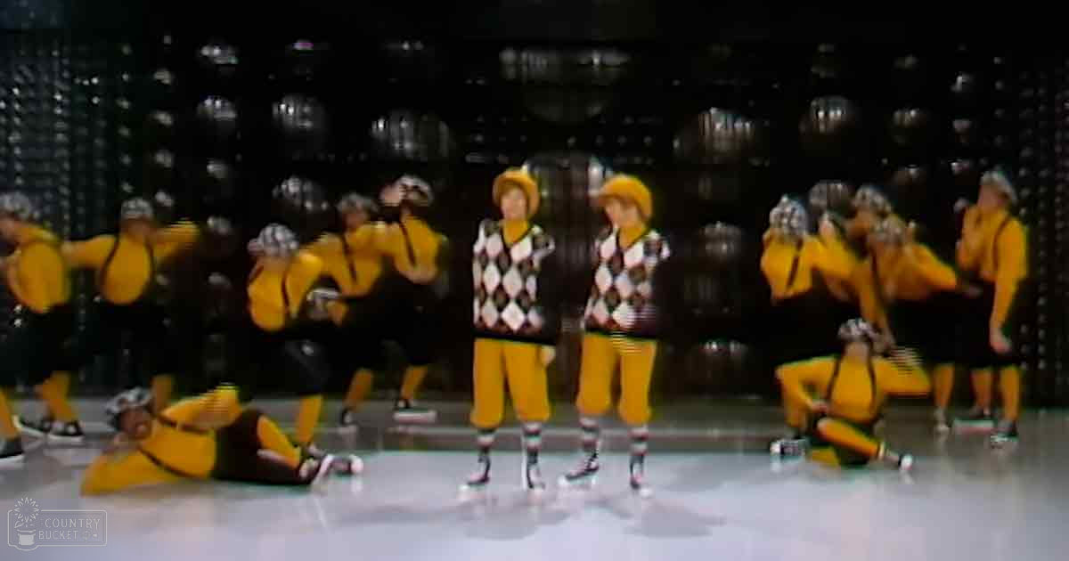 Group performance scene on an indoor stage with young adult male singers wearing argyle-patterned sweaters, bright yellow shirts with black suspenders, and roller skates under artificial lighting.