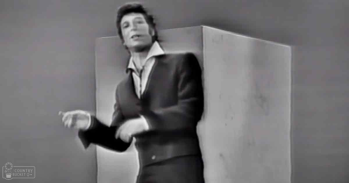 A young adult male wearing a tailored black suit jacket with a white shirt, standing indoors near a worn wooden podium.