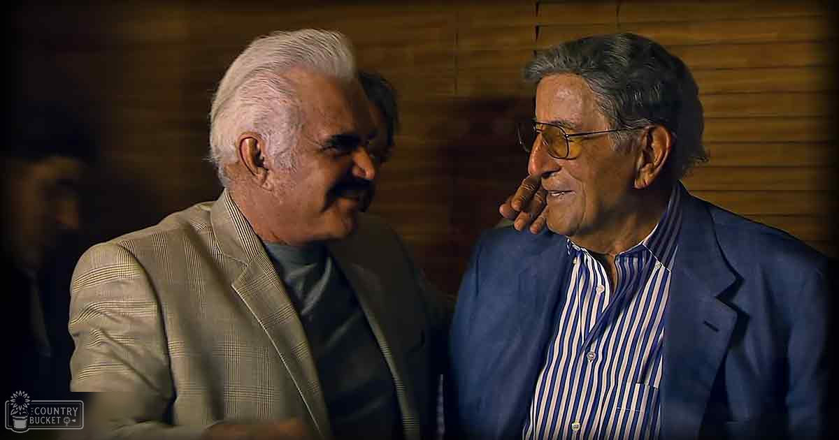 Two older adult males in casual indoor attire, one wearing a beige plaid blazer over a dark gray turtleneck sweater, the other in a blue striped button-down shirt under a navy blue blazer.