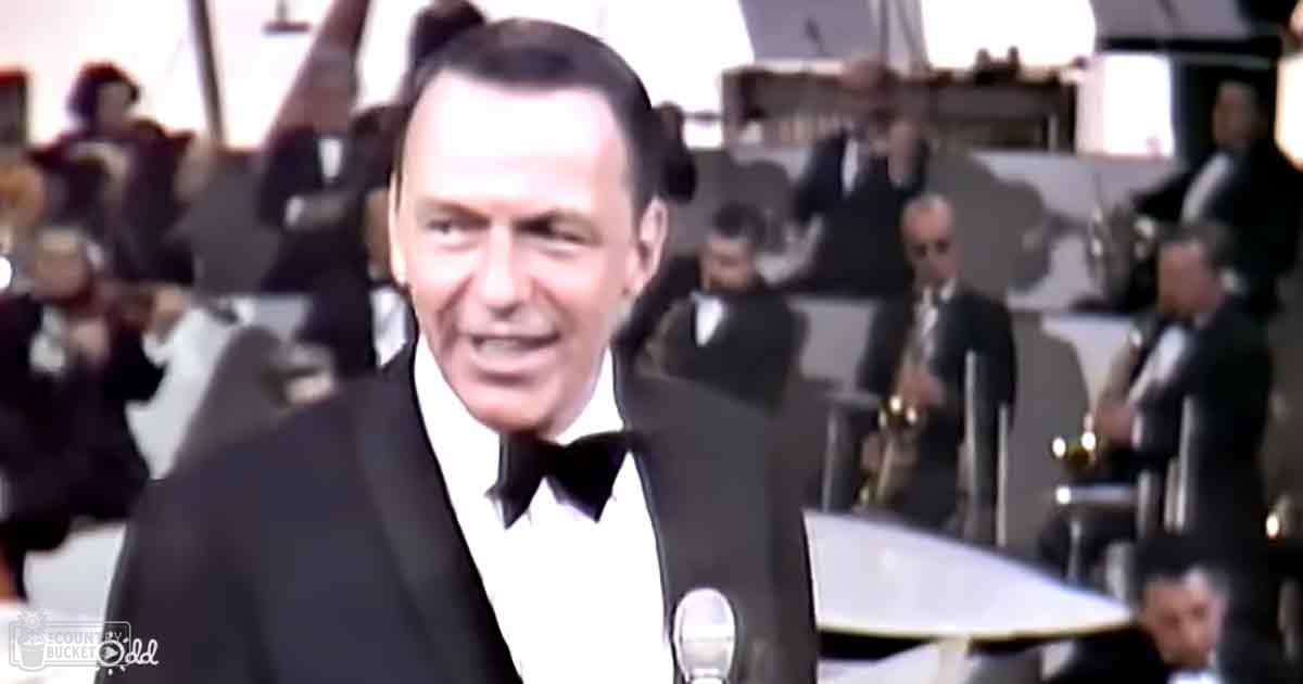 Adult male in his 50s-60s wearing a black tuxedo, performing at an indoor musical event with band setup and microphones.