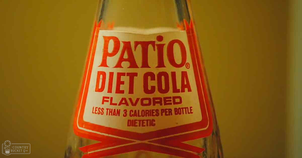 A glass bottle labeled 'Patio Diet Cola' with a red and white label, set against a yellowish wall or surface in an indoor setting.
