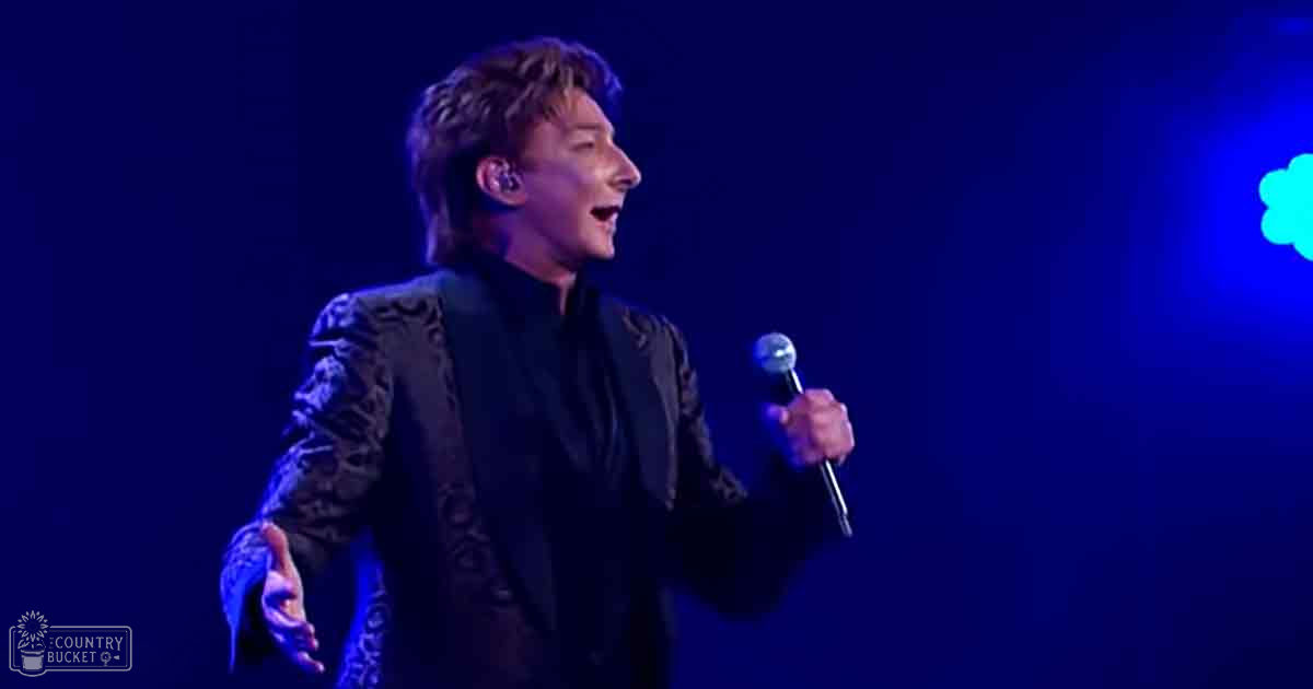 A male adult singer in a dark, patterned velvet suit jacket with intricate embroidery performs on stage under blue lighting.
