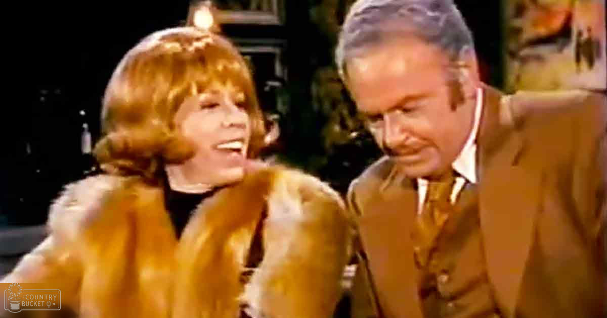 An adult female wearing a golden-brown fur coat with high collar and loose fit stands indoors while an adult male in a brown suit jacket and white shirt is present, the setting appears to be dimly lit and possibly a room or studio.