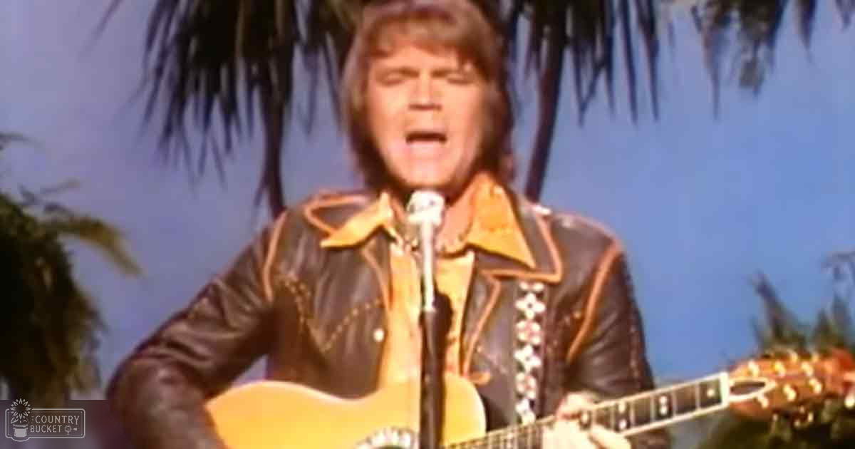 Middle-aged male singer in a black leather jacket with floral embroidery, playing an acoustic guitar under natural daylight against a palm tree backdrop during what appears to be summer or spring.