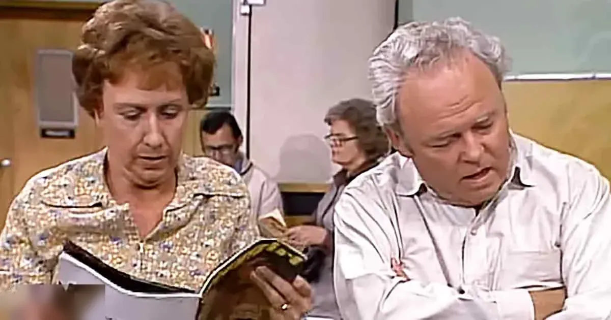 A middle-aged woman wearing a floral-patterned blouse sits at a desk, reading from a book in what appears to be an indoor classroom or office setting with whiteboard and desks visible.