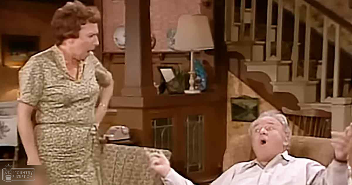A middle-aged woman wearing a floral dress and holding something small in her mouth is seated indoors with an older male adult man who appears surprised on a chair, surrounded by home decor items like framed pictures and a clock. The setting suggests a cozy living room or family room during the daytime.