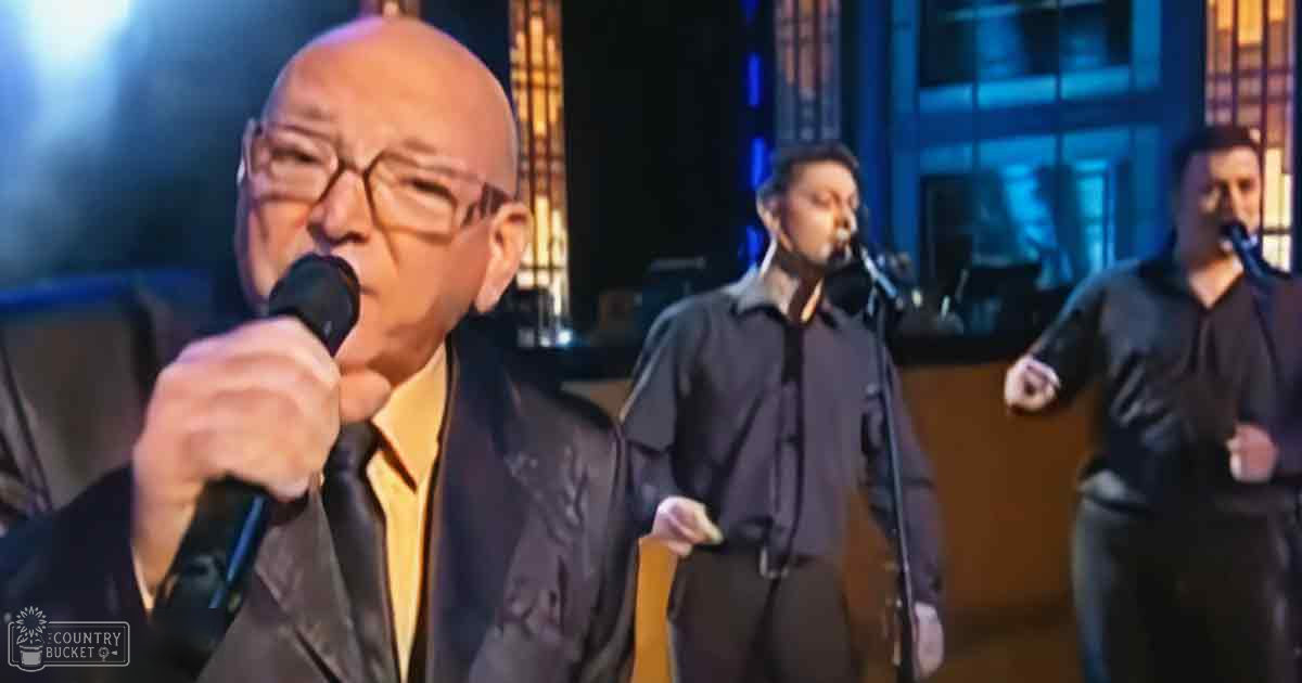 An older adult male singing into a microphone in what seems to be a studio or stage setting, with microphones and controlled lighting creating dramatic effects on performers.